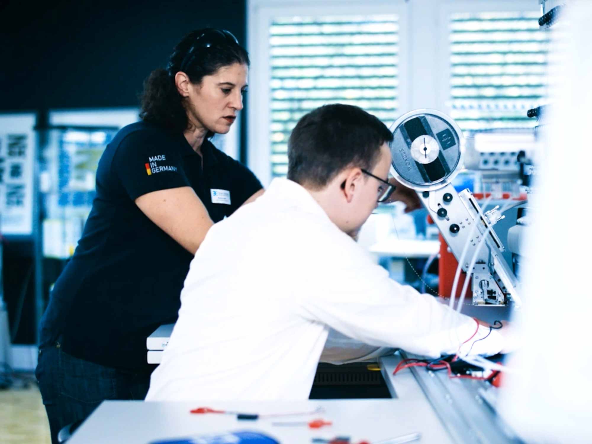 Ein Schulungsteilnehmer und ein Kursleiter arbeiten gemeinsam an einem technischen Sticksystem der ZSK. Bild zweier Menschen, die an einer Technischen Stickmaschine der ZSK arbeiten.